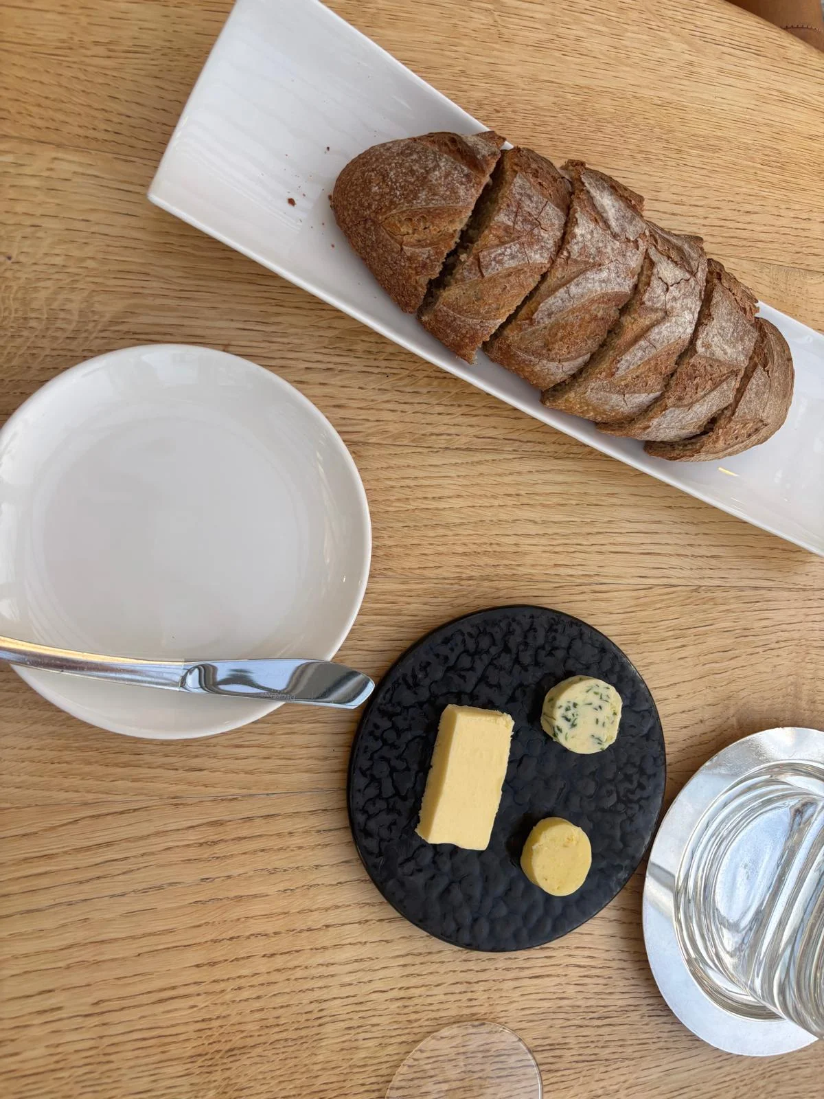 A black slate platter with three Breton butters: a golden classic block, a herb-flavoured round, and a small yellow round, served alongside sliced sourdough