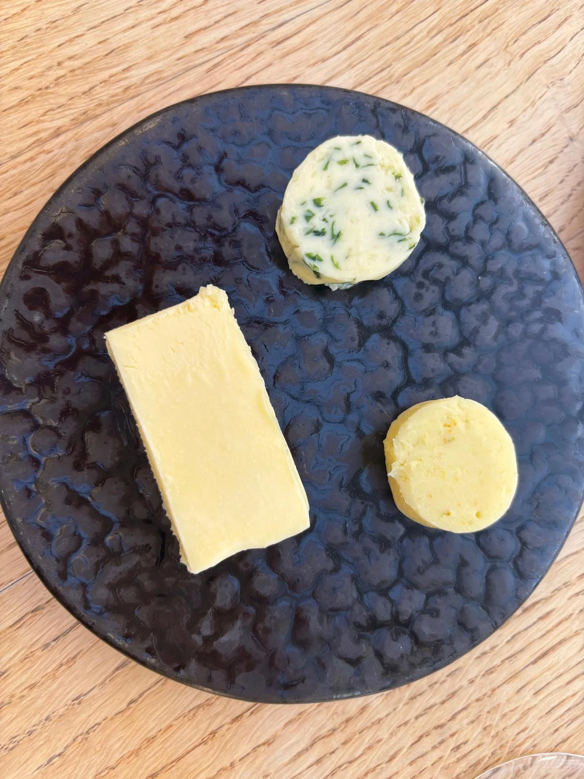 A close-up of three Breton butters on a black slate plate: a yellow salted block, a green herb-flavoured round, and a small golden round