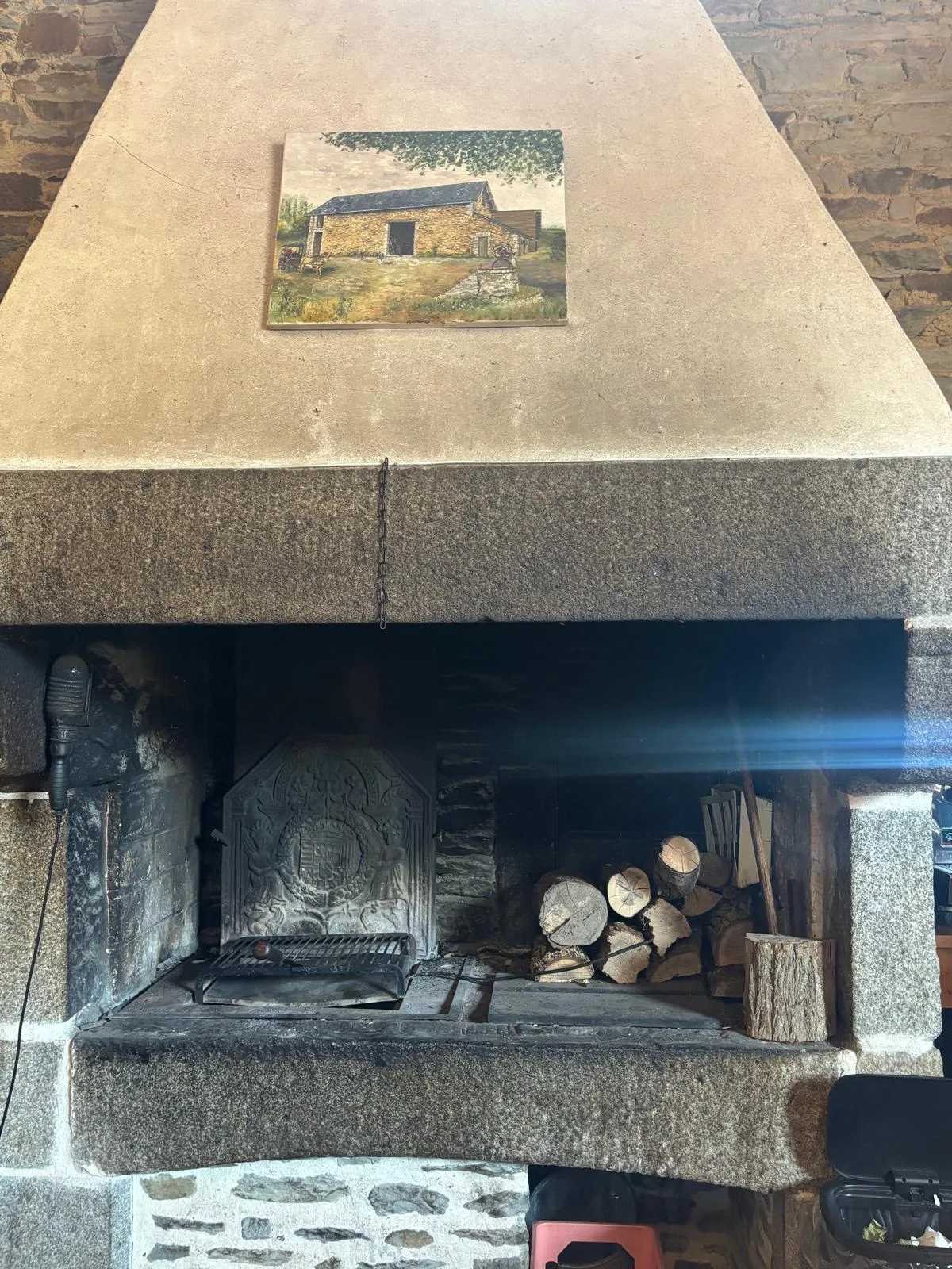 A large stone fireplace at La Ferme Saint-Michel with logs and a painting of the farmhouse above the mantel