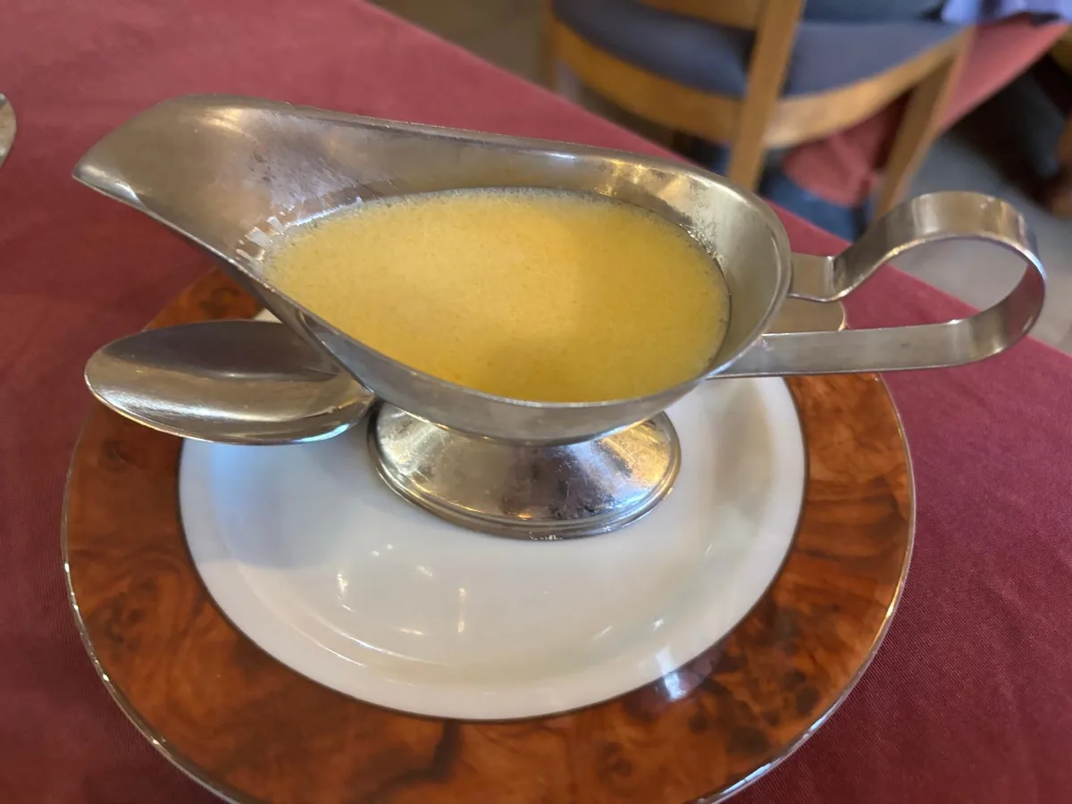 A silver sauciere filled with golden butter sauce on a burl wood saucer