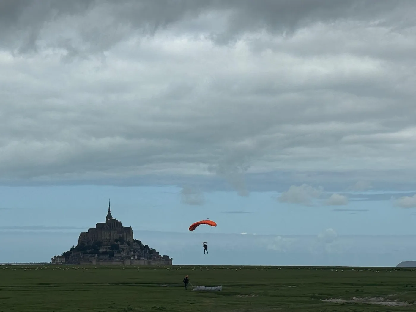 A paratrooper with an orange canopy coming down over the bay beside Mont-Saint-Michel